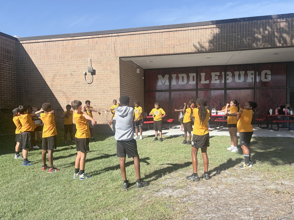 Multiple young individuals in yellow shirts and black shorts are standing on a grassy field in front of a building.