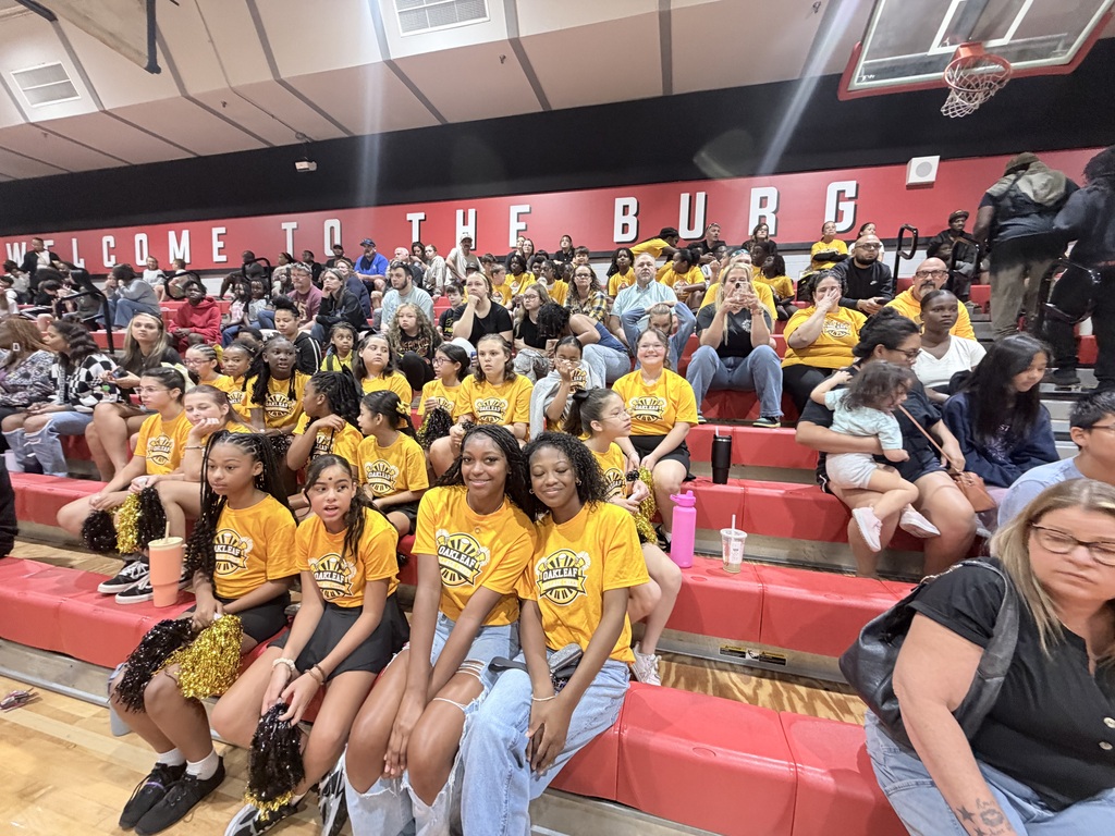 People in yellow shirts and jeans sit on bleachers in a gym. The bleachers are red.