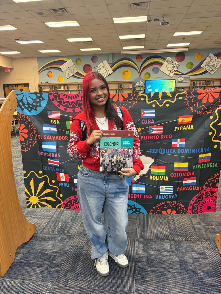 Person in red top and jeans holds a book in front of a colorful backdrop with flags.