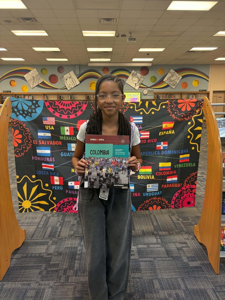 Girl wearing glasses and a black scarf standing in a library, holding a book titled Colombia.