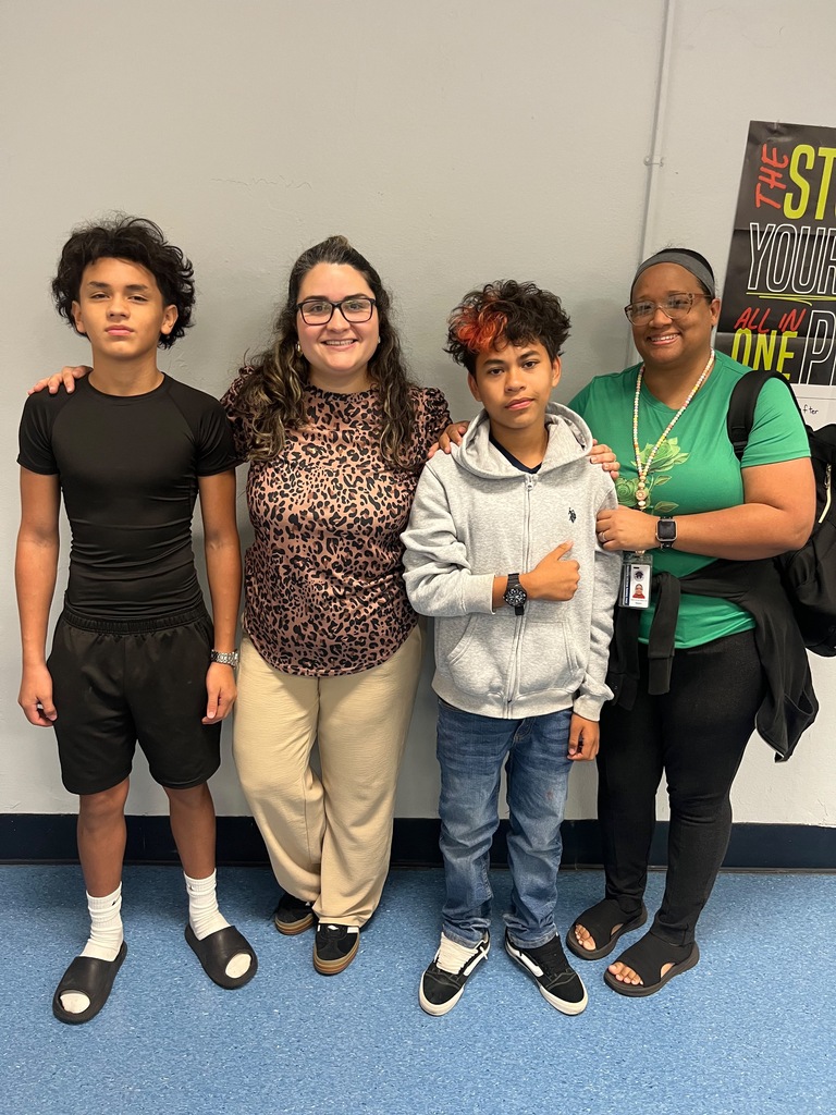 Five individuals stand close together. Two young boys, one in black, the other in gray. Two women, one in leopard print and one in green. A poster is on the wall behind them.