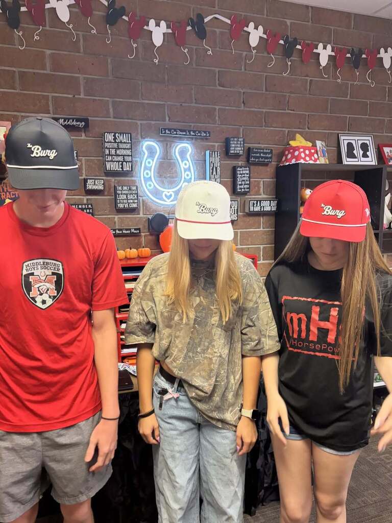 Three people stand in a room. They wear hats. The background features a brick wall and decorative items.