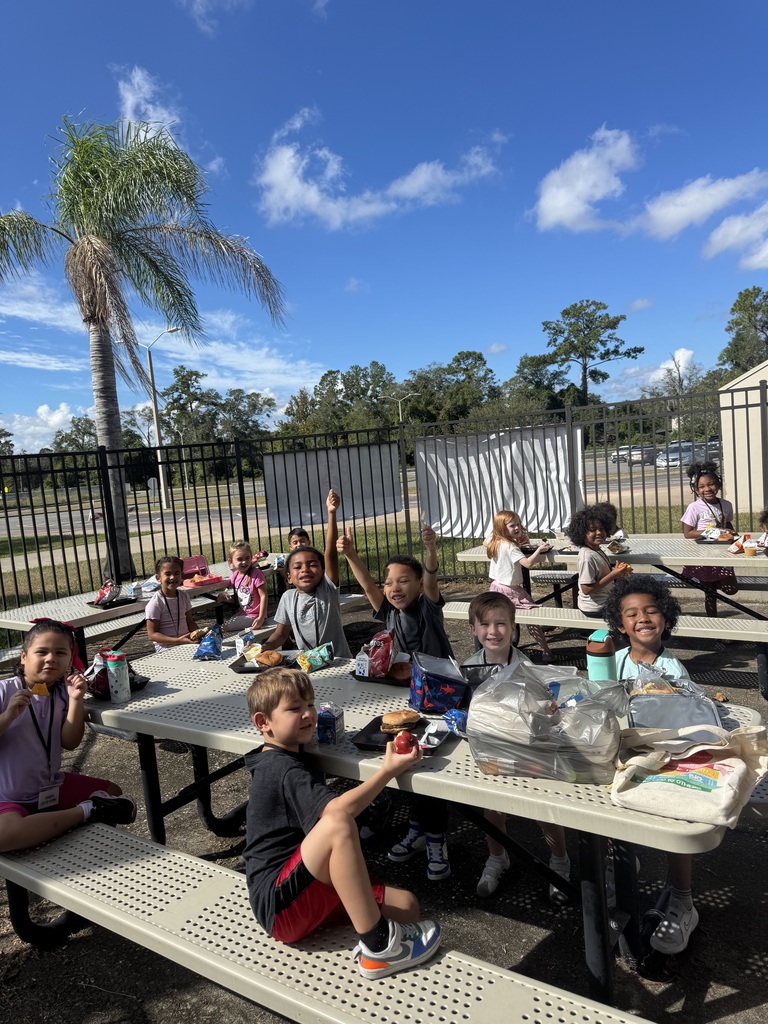 Several children sit at a picnic table under a fence. A few raise their hands. Palm trees and distant trees are in the background.