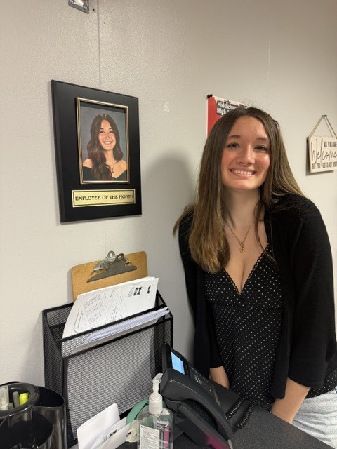 Woman smiling stands at a counter; a photo of a woman is on a wall behind her.