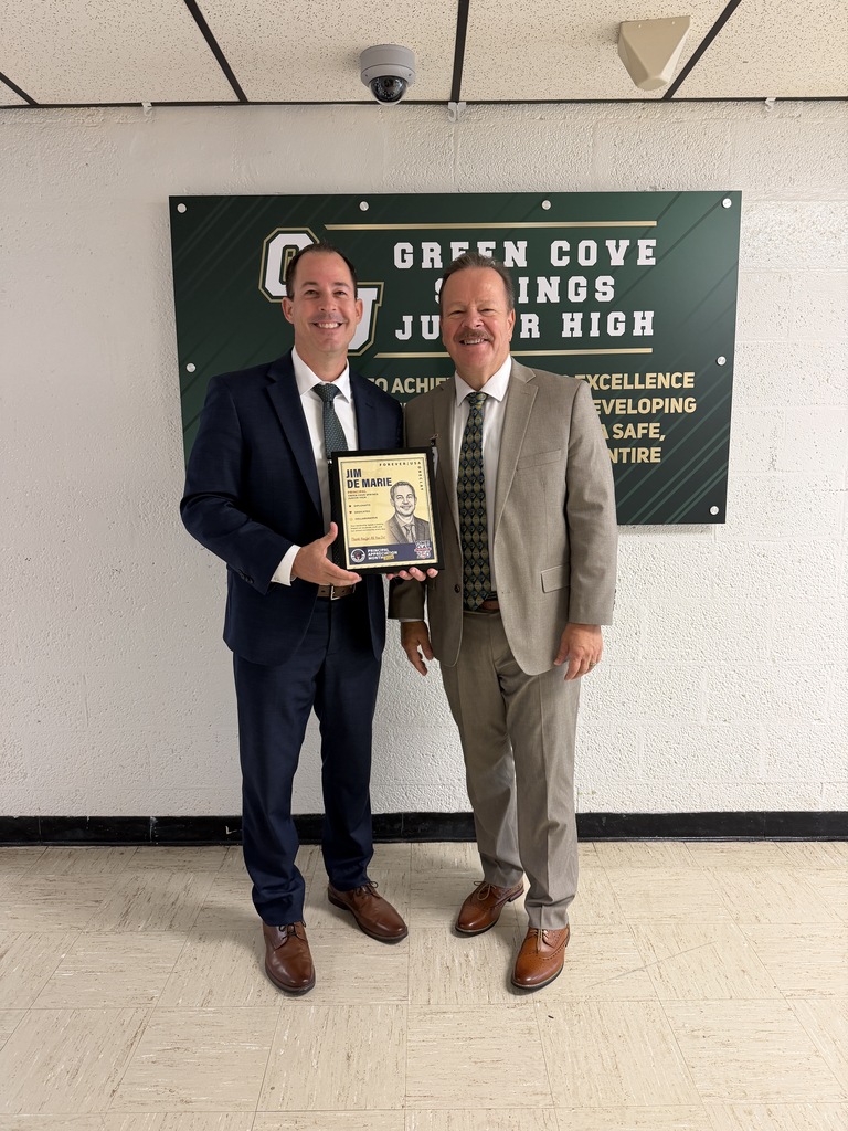 Two men in suits stand before a Green Cove Junior High banner, one holding an award plaque.