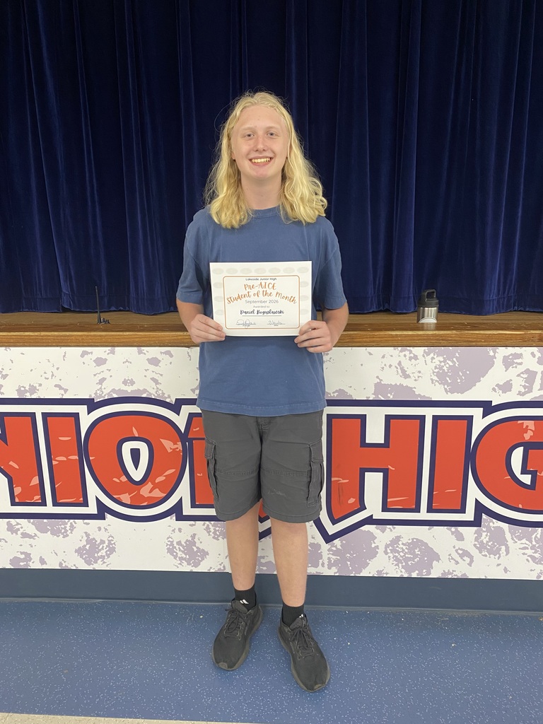 Blonde teen with blue shirt and gray shorts stands holding an award in front of blue curtains.