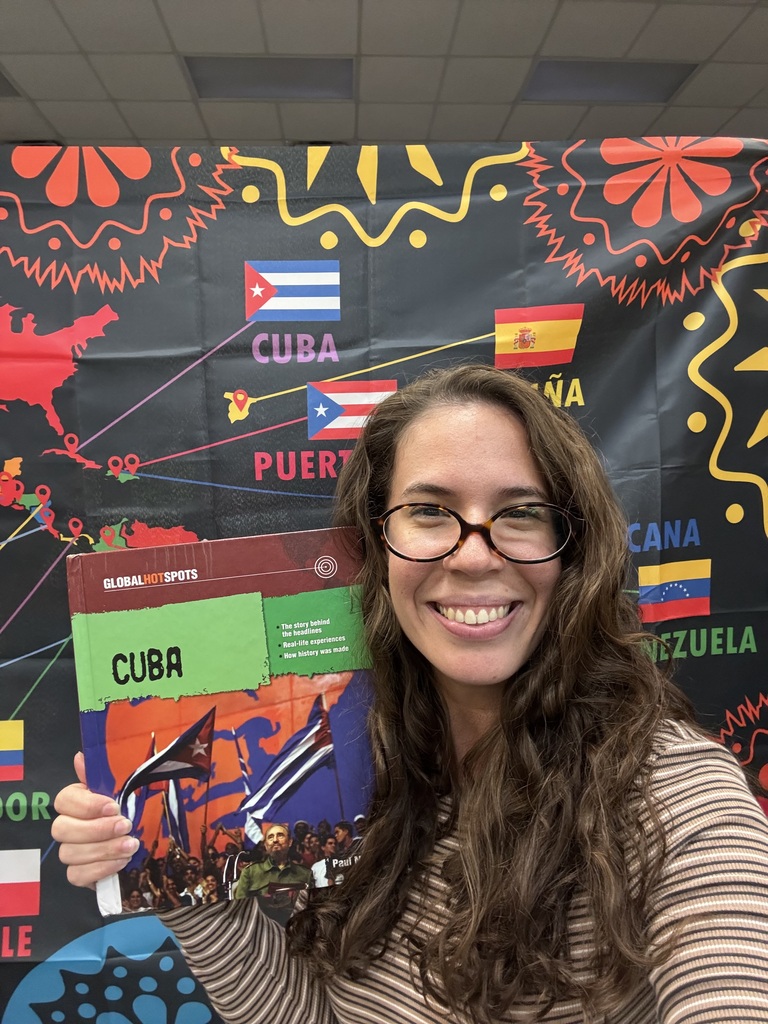 Woman with glasses holds a book titled "CUBA" and smiles. Background features flags of Cuba, Puerto Rico, and other countries.