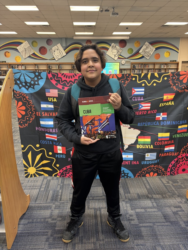 A young person poses with a book in front of a large map displaying countries.