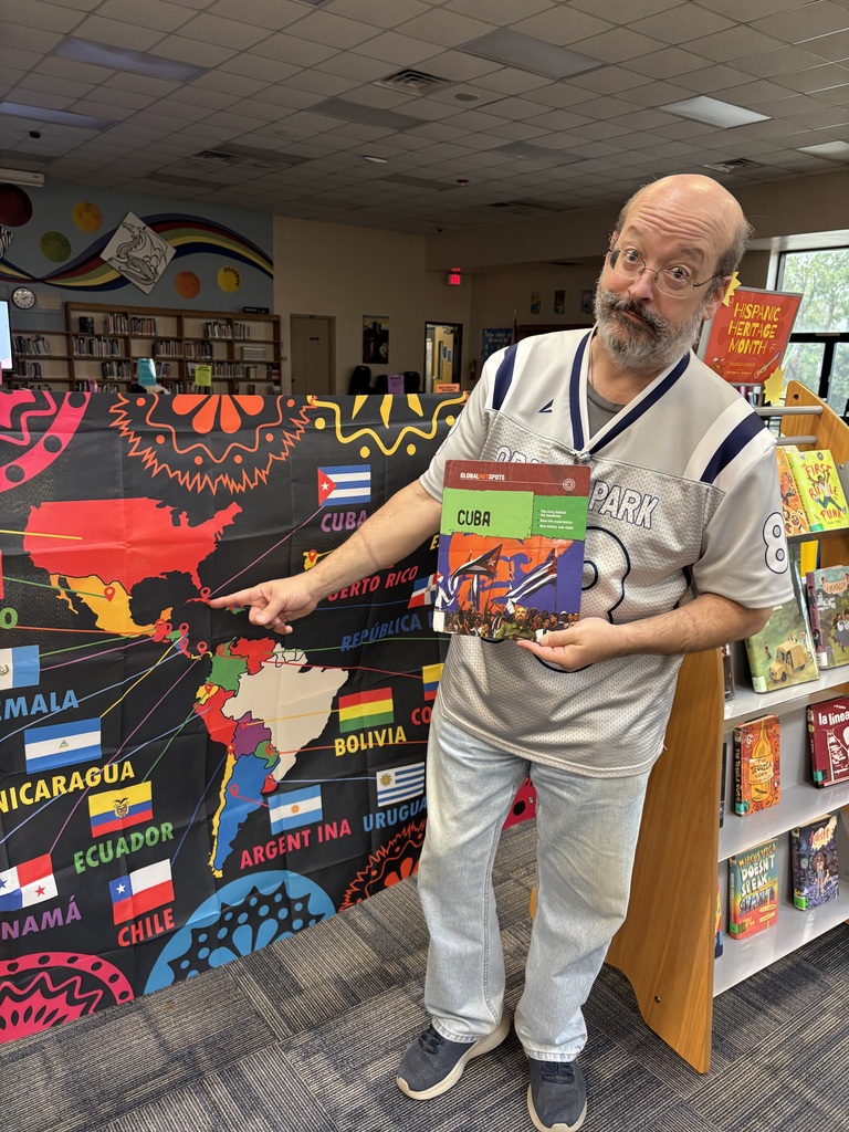 A man wearing glasses and a polo shirt stands before a colorful world map, holding a book in his hands.