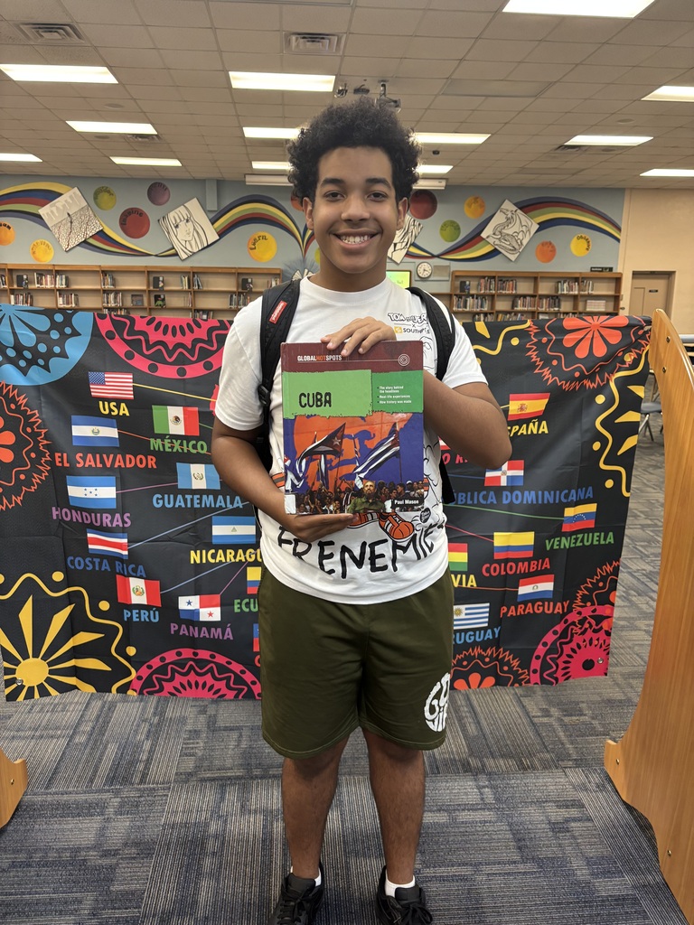 A boy in a library holds up a book titled "Pernemer" against a colorful backdrop with flags.