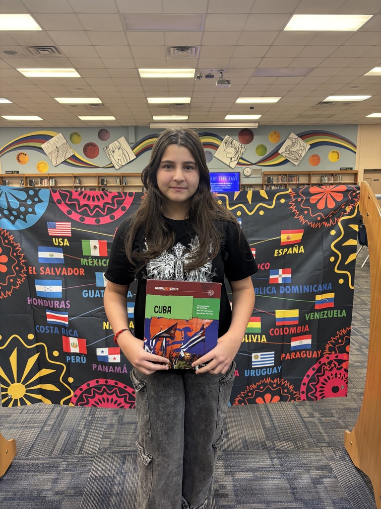 Girl in black t-shirt holding a book, standing in front of a library backdrop with flags.
