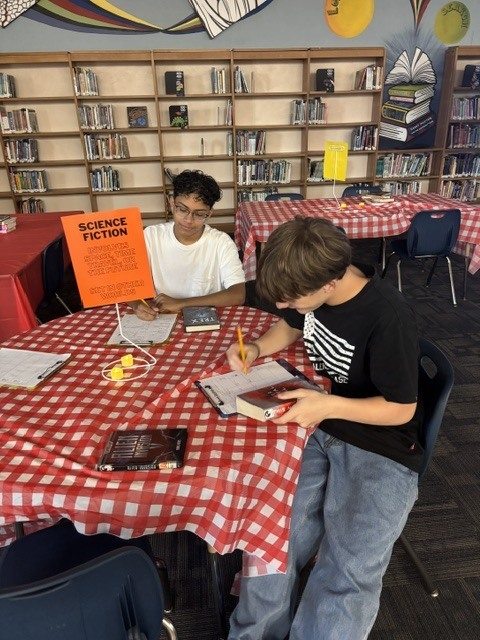 Two boys write at a table in a library. One is in a black shirt, the other in white. Books and papers are on the table.