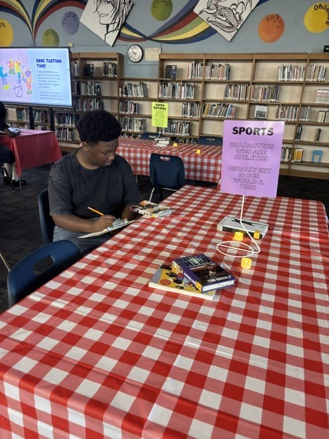 Person sitting at a red-and-white checkered tablecloth, with books and a sign that says "Sports." Behind is a library with wooden shelves and a projector screen.