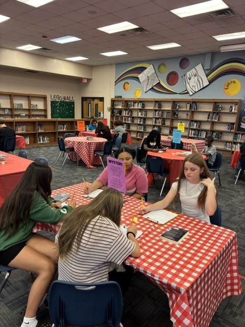 Group of people sitting at tables with red and white tablecloths in a library. Bookshelves in the background.