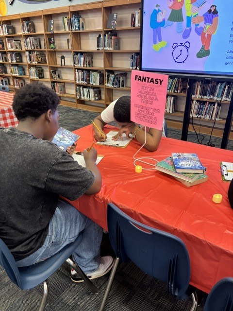 Two people seated at a table, one writing, with a fantasy sign in the background.
