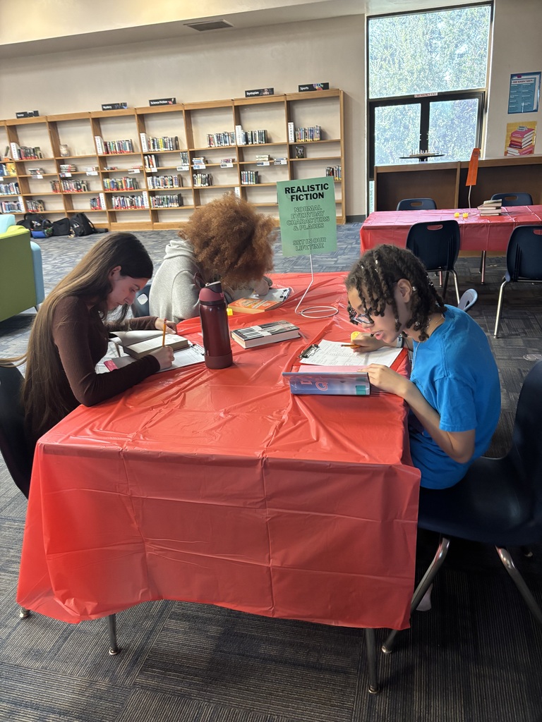 Three people sit around a table, writing in books. A table has a red cover and books on it. Shelves in the background are filled with books.