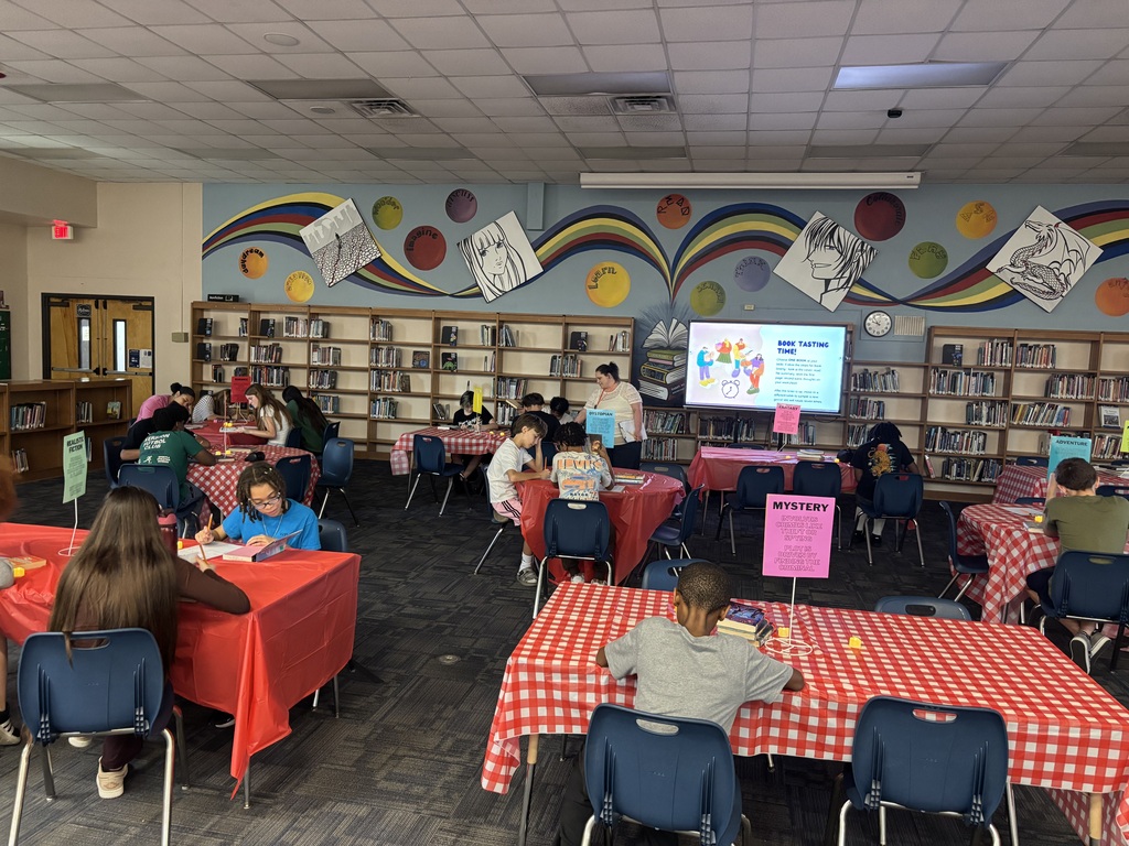 Library room with students sitting at tables. Colorful designs adorn the walls. A screen is mounted on the wall.