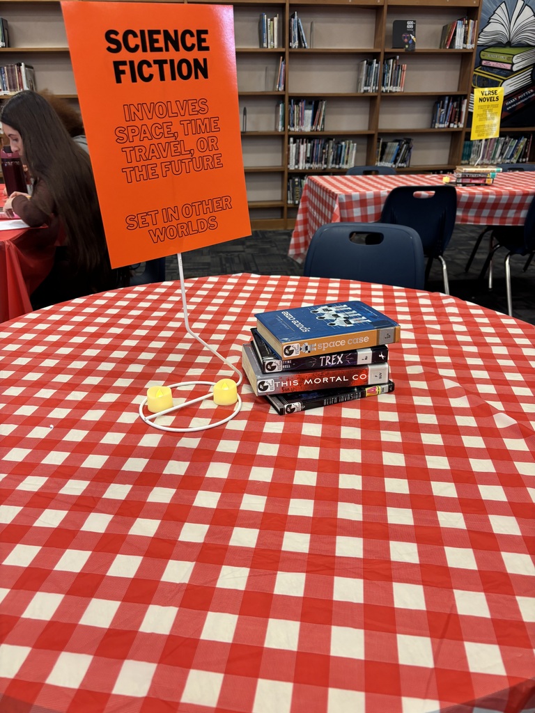 A table with a red-and-white checkered tablecloth holds books and a Science Fiction sign. Behind it, a library features bookshelves with books.