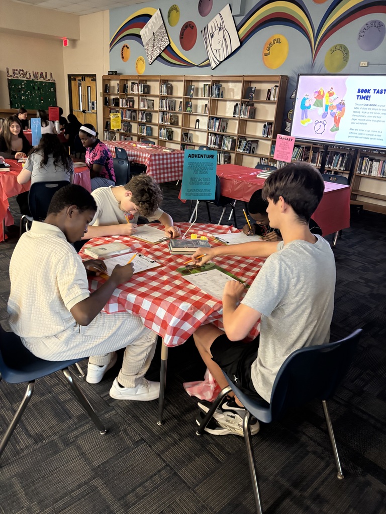 A group of students are sitting at a table in a library with a projector screen on the wall.
