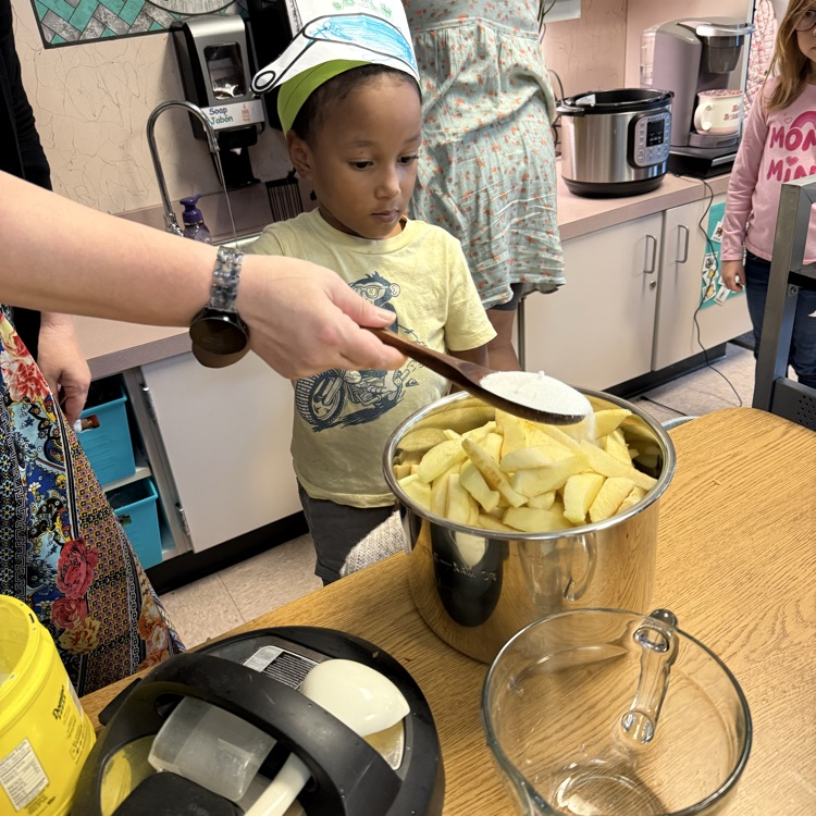 child stands behind pot of apples while an adult pours sugar over them