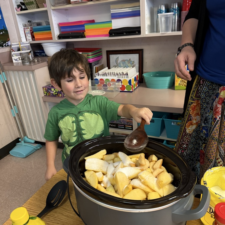 Boy stands behind a pot of apples holding a large wooden spoon