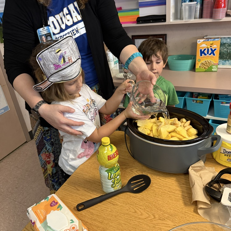 A boy and girl pour water over a pot of apples with the help of the teacher.
