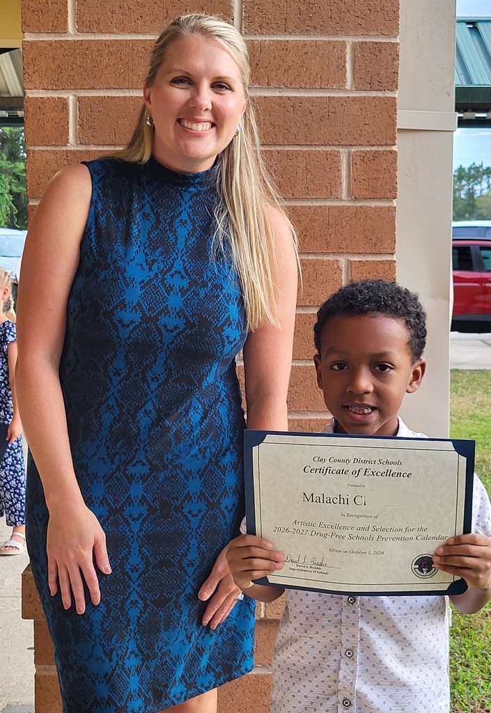 Woman in blue dress and boy in white shirt stand together, holding a certificate. Boy smiles, woman smiles. Behind them is a brick wall and a red vehicle.
