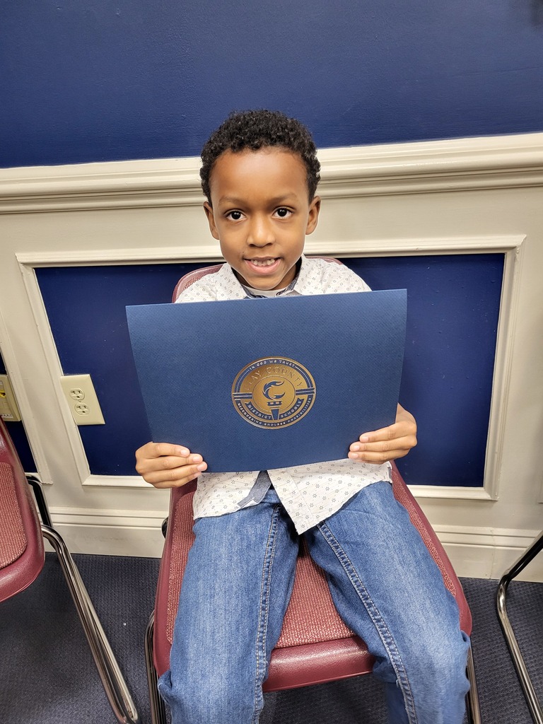 Child holding blue folder with logo, seated in red chair, smiling, looking at camera.