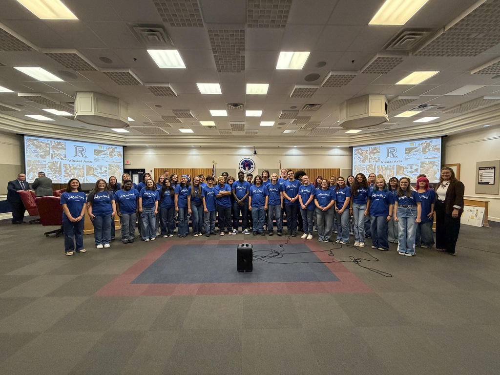 A group of people in blue shirts and jeans stands in a circle around a black object on the floor. Behind them are two large screens.