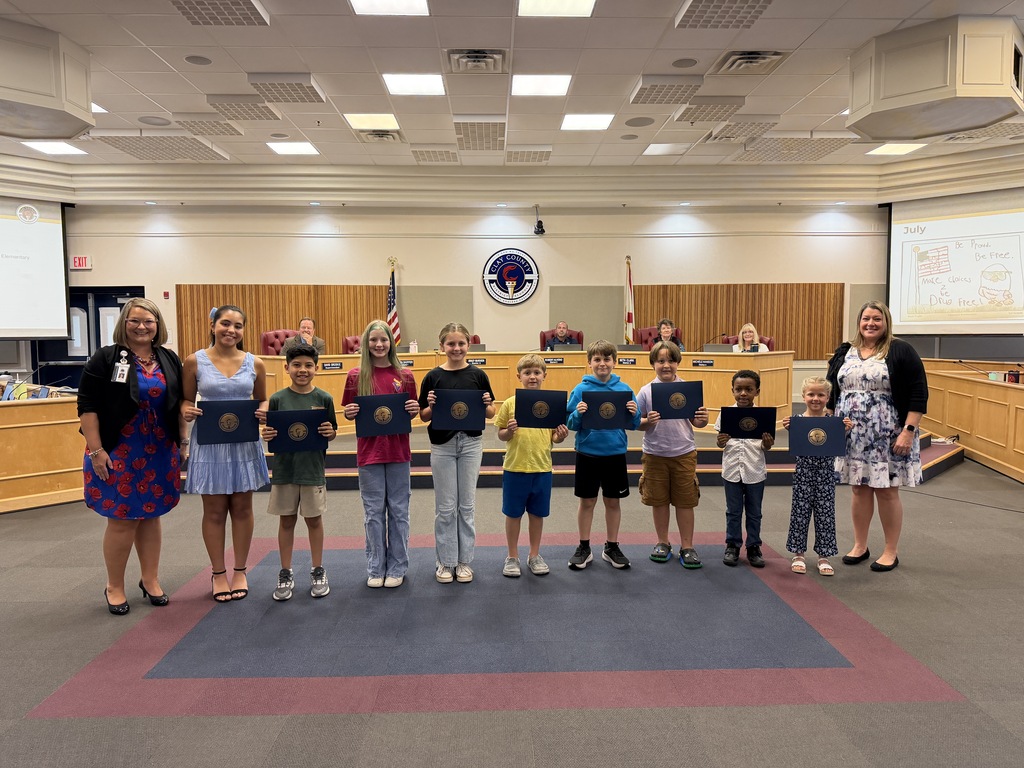 Children and adults stand in a row holding certificates. A stage is in the background.