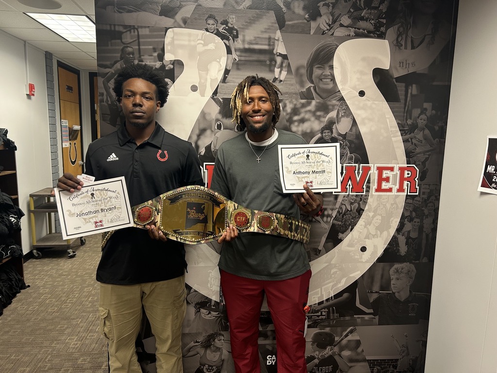 Two men stand next to each other, holding a belt and certificates. They pose in a room with a large mural in the background.