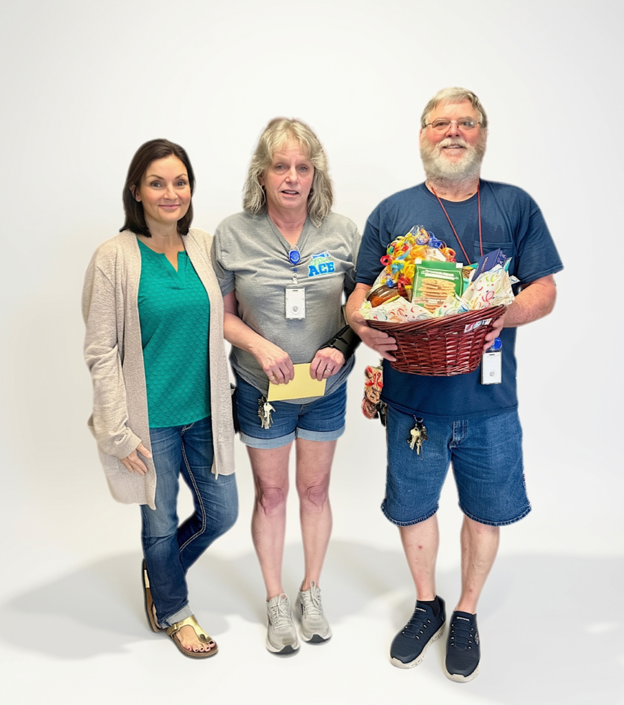 Three people stand together, one holding a basket of food. The woman wears a green shirt and denim shorts. The man holds the basket.