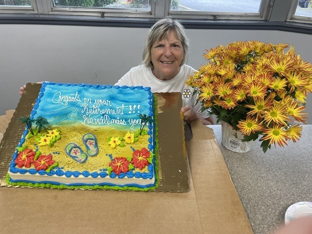 A woman sitting at a table holds a cake decorated with a beach scene and flowers.