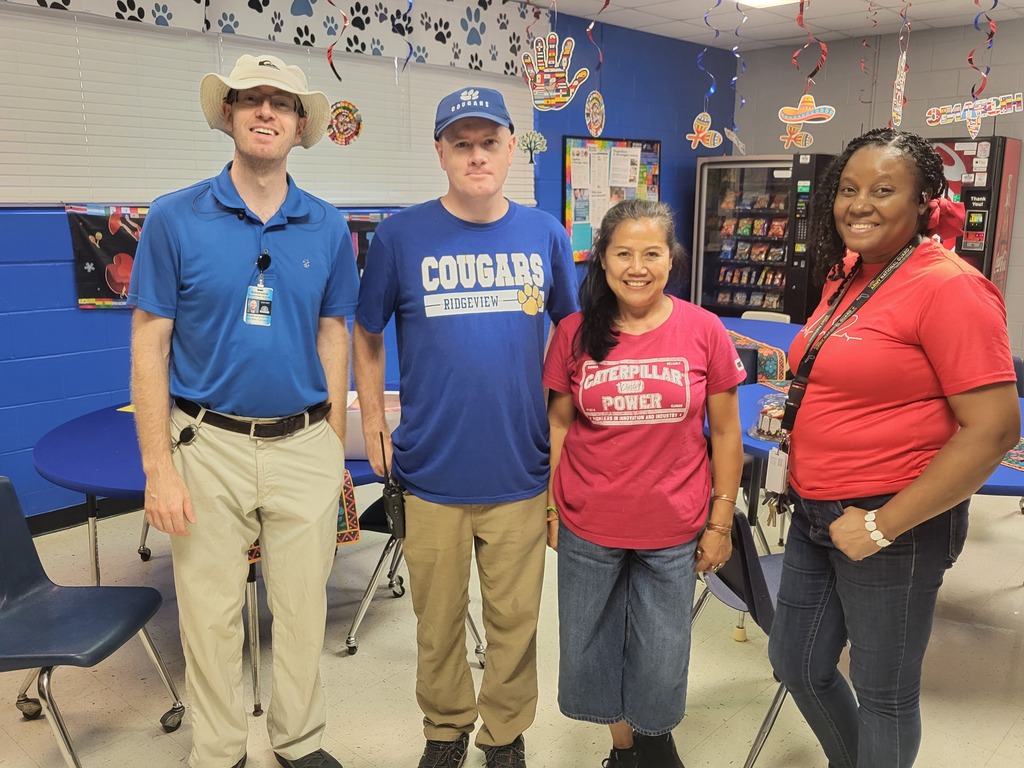 Four people standing in a classroom. Two men and two women. The woman on the right wears a red shirt.
