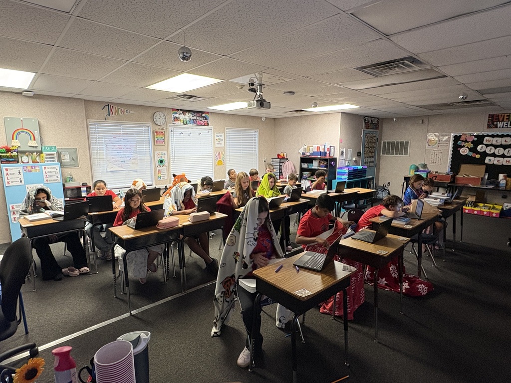 A classroom with students seated at desks, using laptops, and a teacher standing. Desks are arranged in rows.
