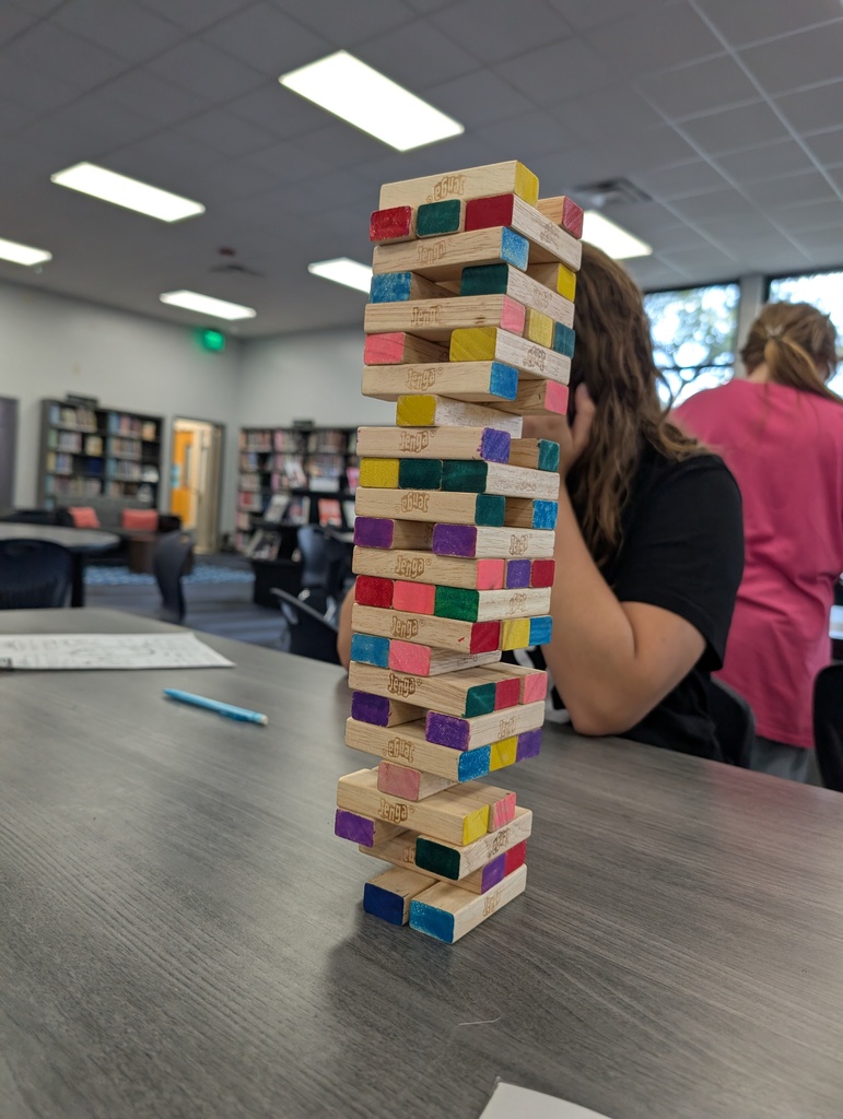 A tall stack of colorful Jenga blocks stands on a table in a library. A person sits nearby.