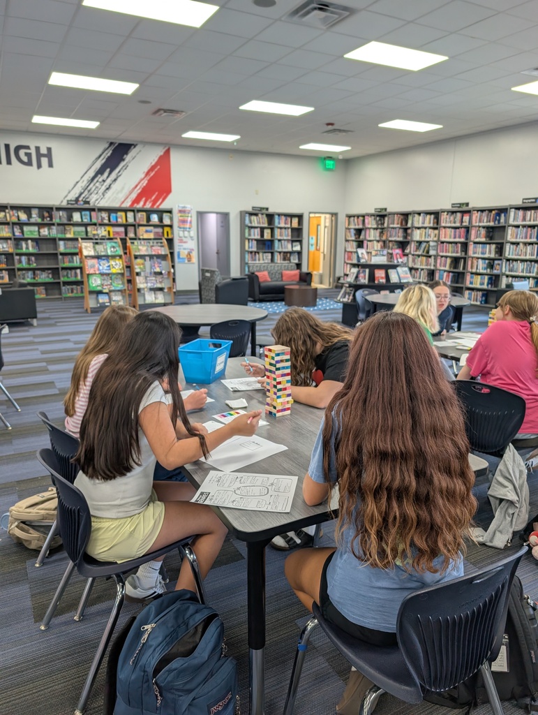 Library tables with students seated. One student stacks blocks. One student has a backpack.