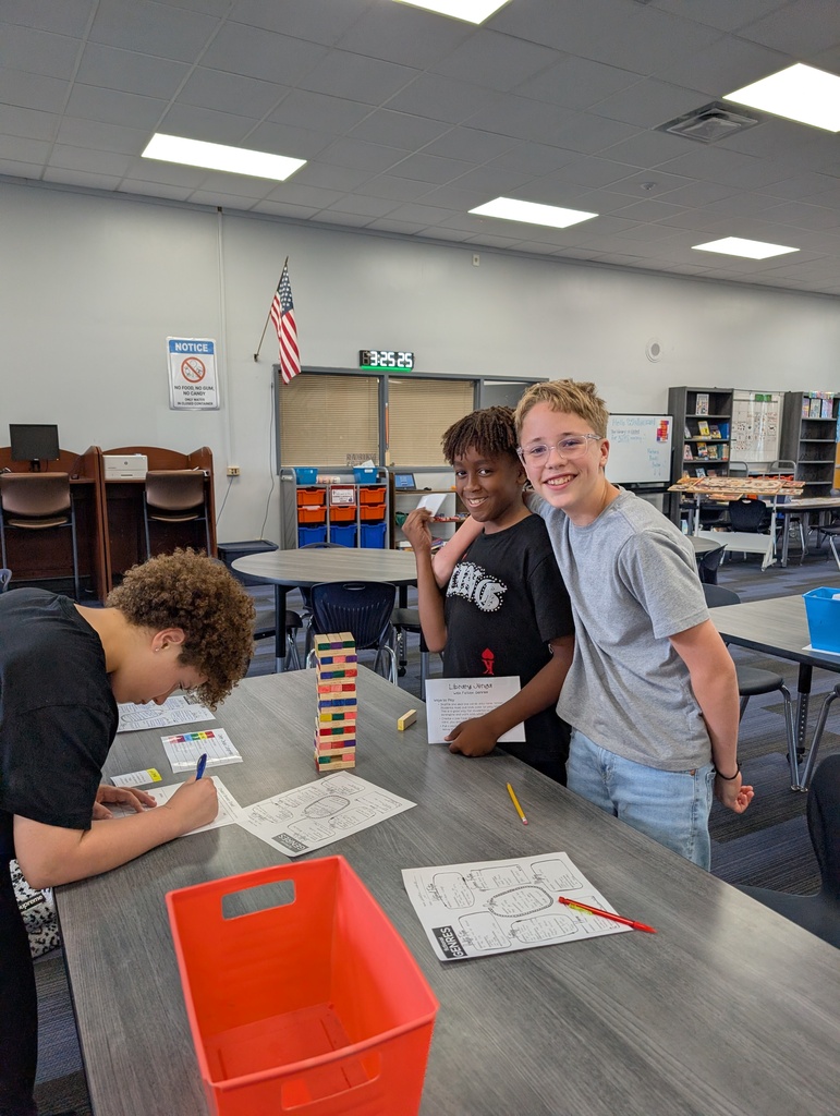 Three young people stand around a table, with one focused on writing on paper. A flag and bookshelves are in the background.