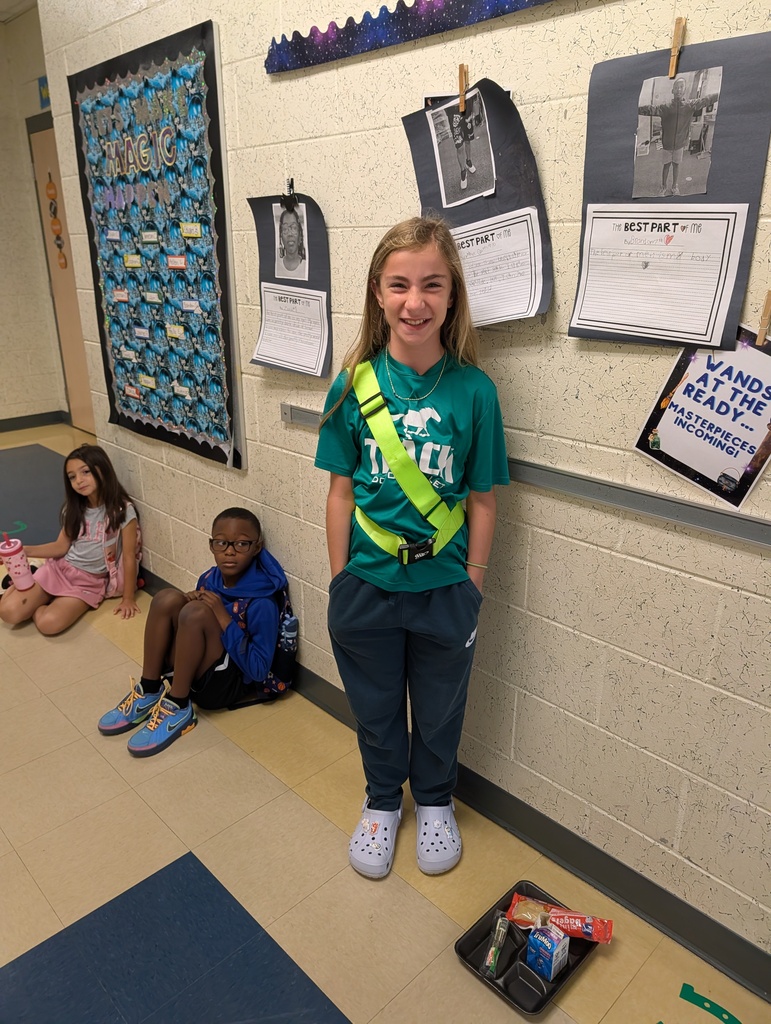 A girl in a green shirt and reflective belt stands in a hallway. Two children sit on the floor behind her. Bulletin boards are on the wall.