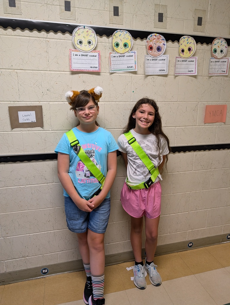 Two young girls wearing reflective belts stand next to each other in front of a wall with paper circles.