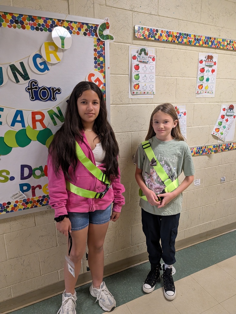 Two girls in safety gear standing near a classroom wall. One has a pink jacket and denim shorts, the other a gray t-shirt and black pants.
