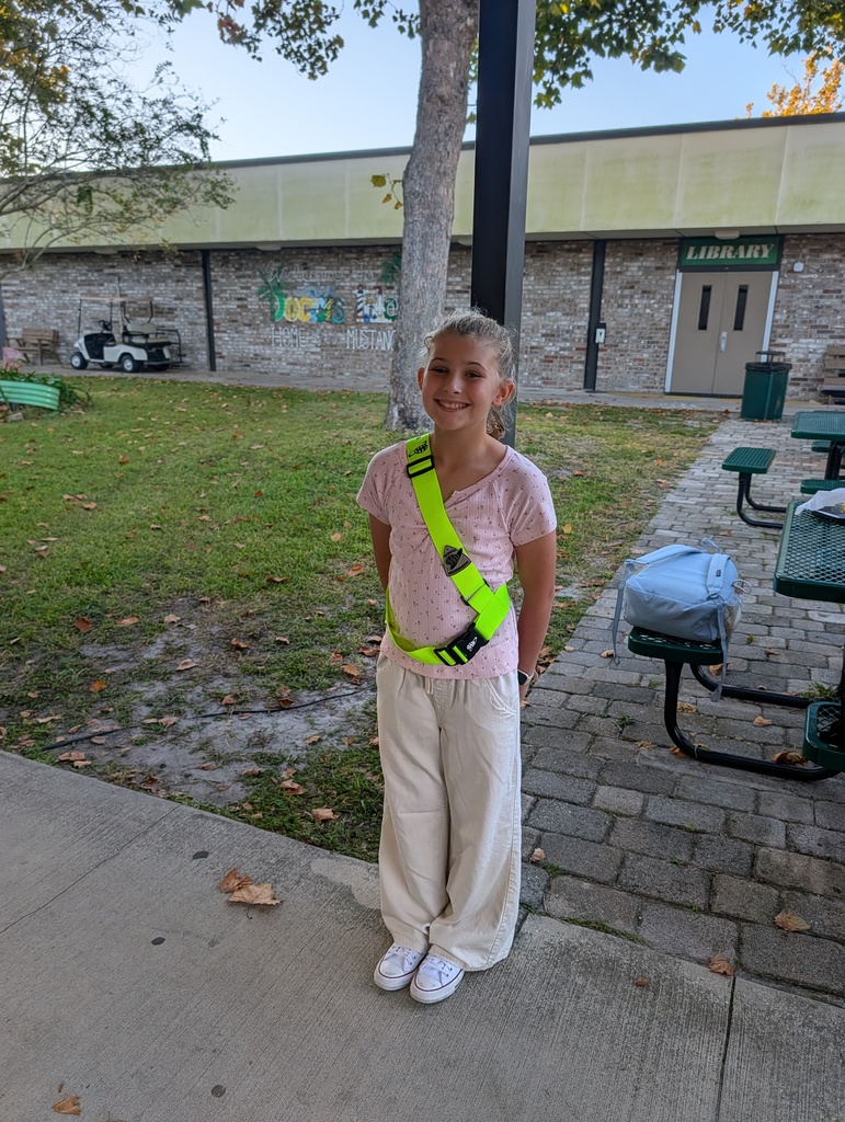A young girl wearing a reflective safety vest and white pants stands on a concrete sidewalk. Behind her, there is a brick pathway, tables, and a building.