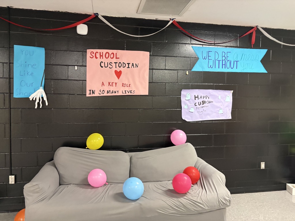 A gray couch with balloons of various colors. Behind it, a black wall displays signs that read "SCHOOL CUSTODIAN" and "WE'RE WITHOUT."