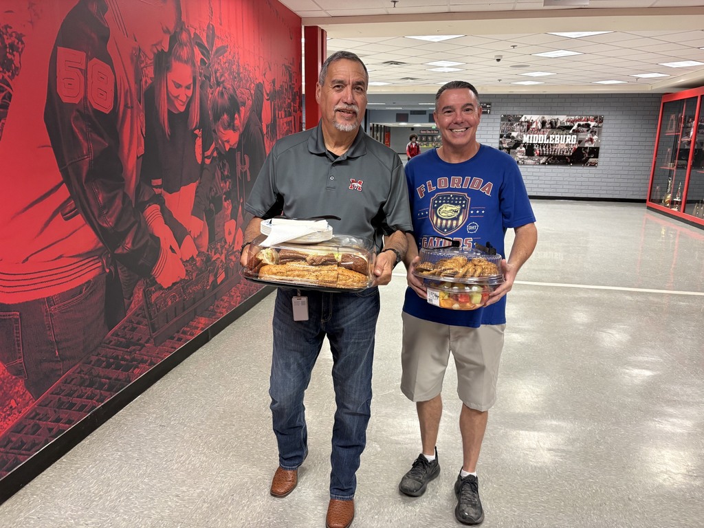 Two men stand in a hallway, each holding a plate of food, with a red wall mural behind them.