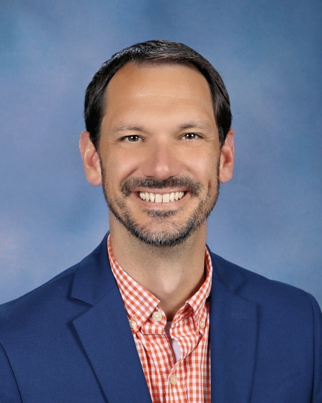 A man smiling with a beard, wearing a blue blazer and a red checkered shirt against a blue background.
