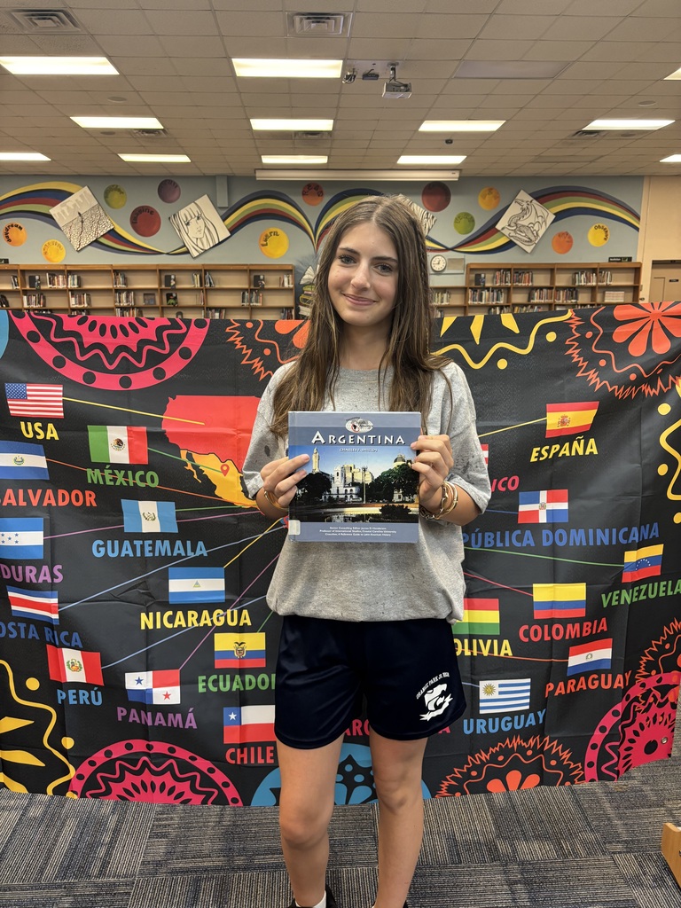 A woman stands in front of a colorful backdrop with flags from various countries, holding a book.