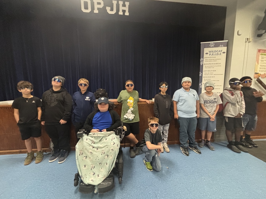 A group of people in casual attire, some in sunglasses, stand behind a stage with "OPJH" on a blue curtain.