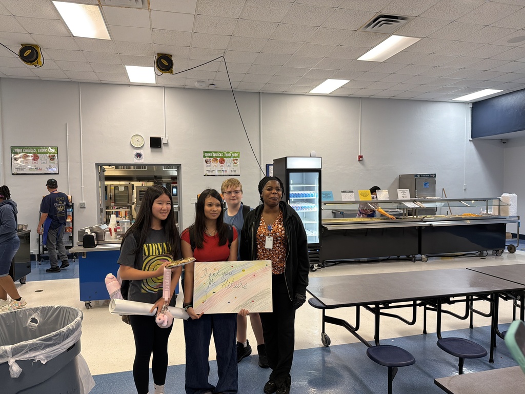 Four students stand in a cafeteria, holding a sign. Behind them are tables and chairs, a trash can, and a refrigerator.