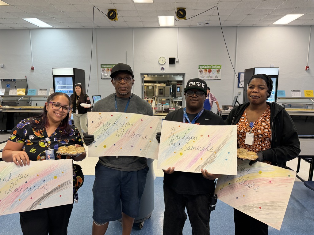Four people in a cafeteria holding up colorful artwork. A woman with glasses stands on the left.