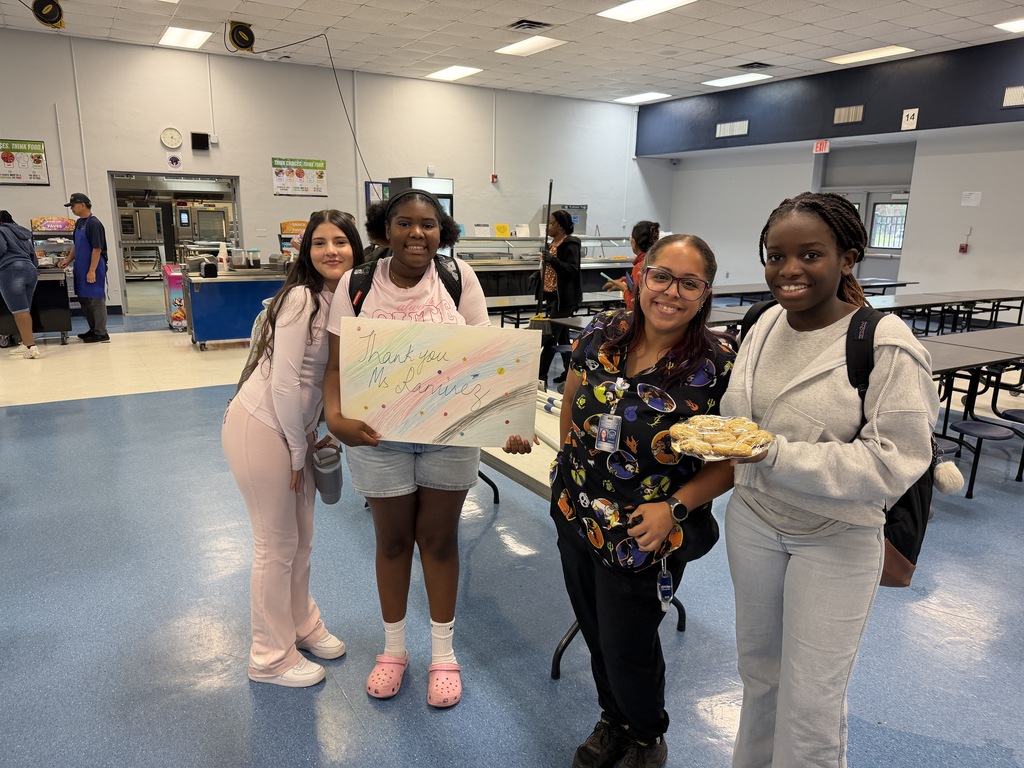 Four women pose in a cafeteria with one holding a sign and another holding a plate.